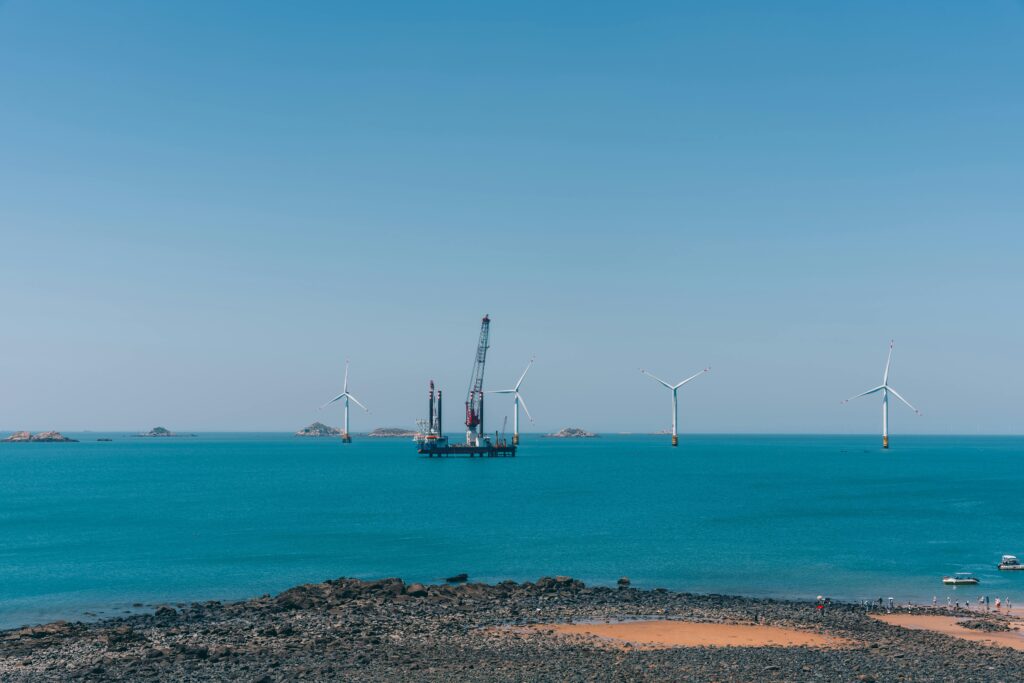Photograph of offshore wind farm, taken from the stoney shore. Clear blue skies and water. 