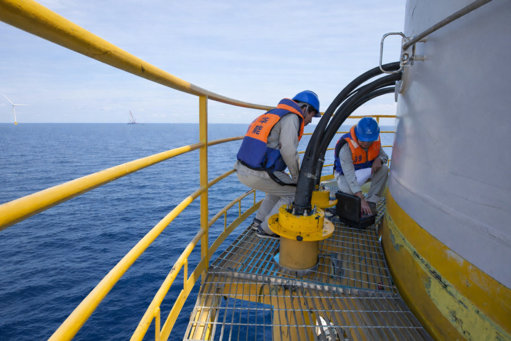 Two offshore technicians in safety helmets and life vests inspect monitoring equipment on an offshore wind turbine platform